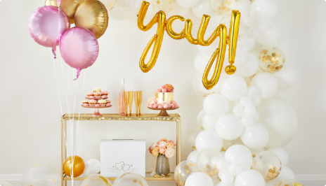 Pink and gold mylar balloons, a gold balloon banner spelling “Yay” and a large white balloon arch surround a gold table s