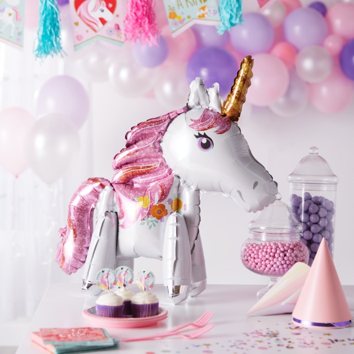 Unicorn shaped mylar balloon on a table with a plate of cupcakes, candy and party supplies.