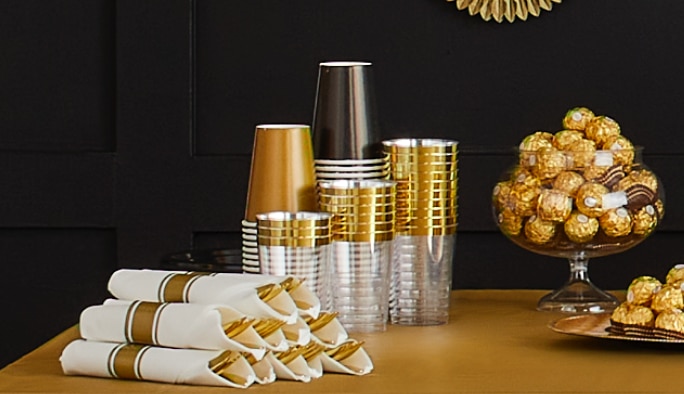 Elegant paper and plastic cups stacked on a tabletop beside rolled napkins and a bowl of chocolates