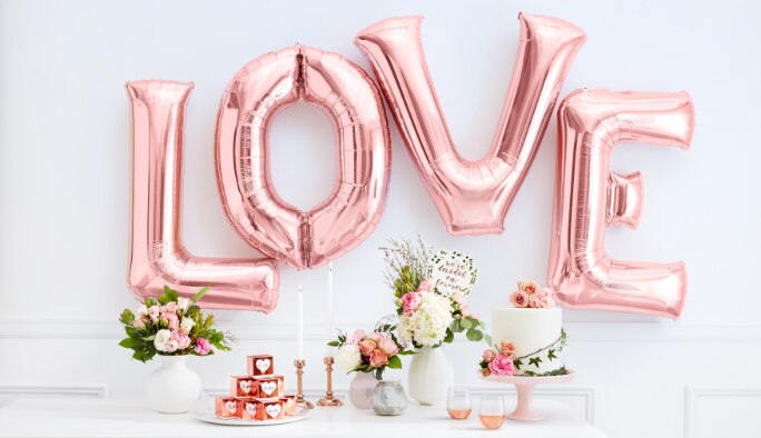 Des ballons or rose en lettres L-O-V-E sont suspendus sur un mur par-dessus une table remplie de fleurs, d’un gâteau et d’une pile de boîtes pour cadeaux-surprises.