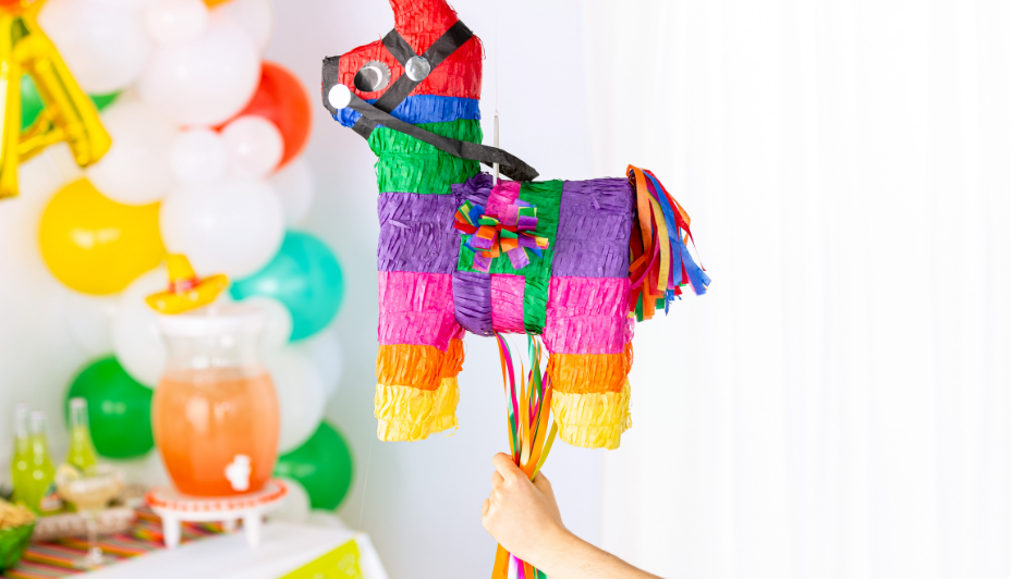 A child’s hand holding a rainbow-coloured jumbo donkey piñata with a variety of balloons and party snacks in the background.