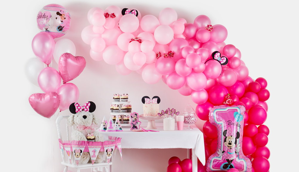 A pink Minnie Mouse-themed balloon arch hangs on a wall next to a dessert table and a highchair decorated with Minnie Mouse accessories. 