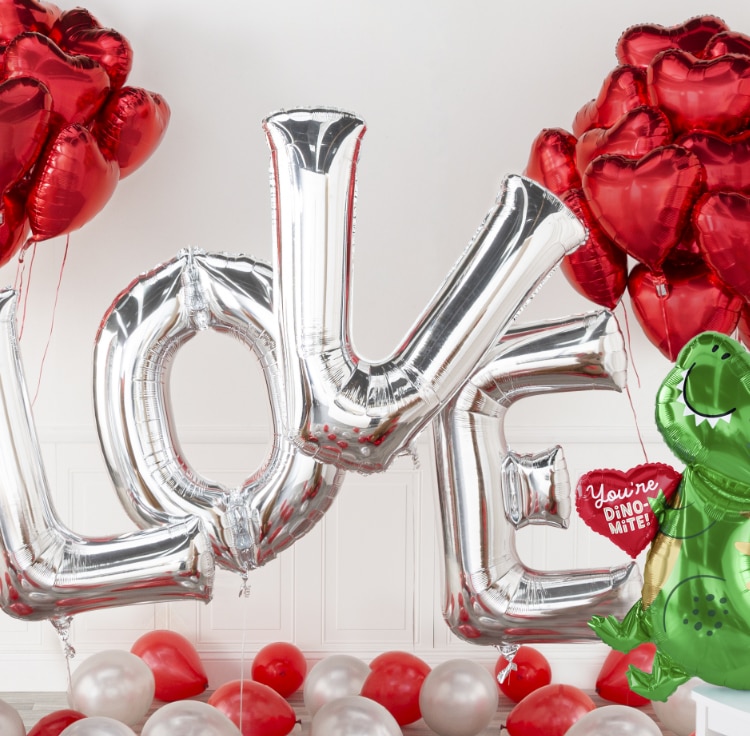 A room filled with silver L-O-V-E letter balloons, a cluster of red heart foil balloons, a You’re Dino-Mite dinosaur-themed balloon.