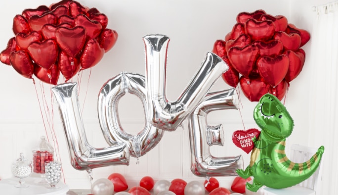 A room filled with silver L-O-V-E letter balloons, a red heart foil balloons, a You’re Dino-Mite dinosaur balloon.