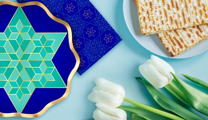 A blue and gold Sophistiplate Passover foil appetizer bowl and napkin on table next to a plate of Matzahs and white tulips.