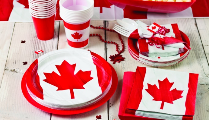 Piles d’assiettes, de serviettes en papier, de verres et de cadeaux-surprises à thème de drapeau canadien sur une table blanche rustique.