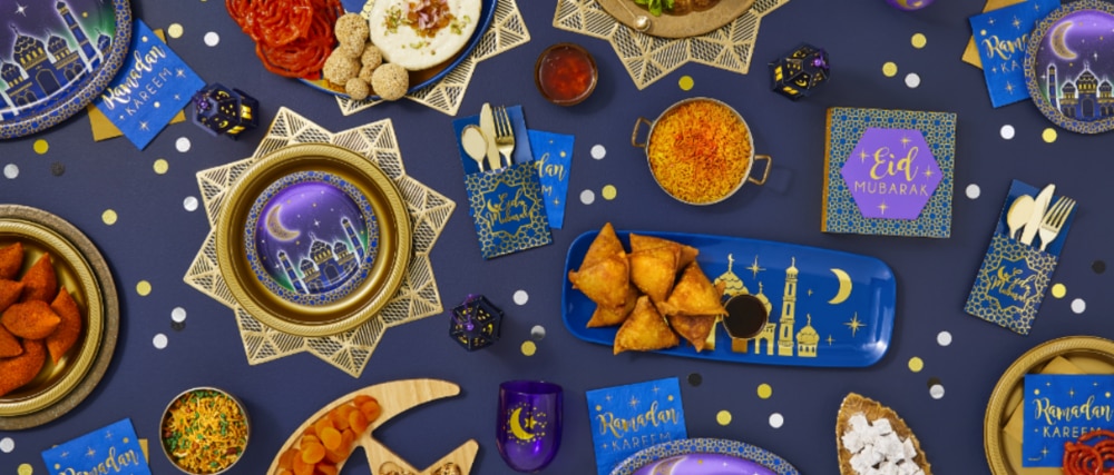 A tabletop filled with Eid dessert plates, an Eid Celebration Long Plastic Platter, and various themed tableware and décor. 