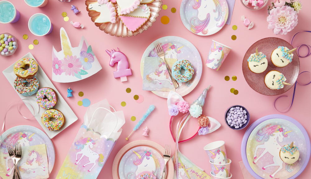 An assortment of Enchanted Unicorn party supplies including a deluxe headband, scattered on a table with confetti and treats.