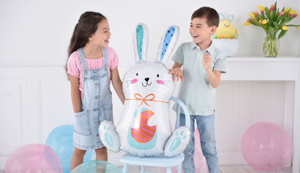Two children stand next to a foil Easter Funny Bunny balloon sitting on a chair.