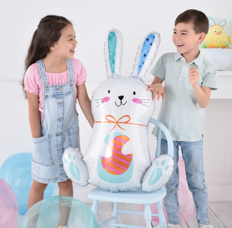 Two children stand next to a foil Easter Funny Bunny balloon sitting on a chair.