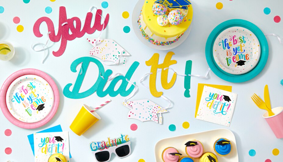 Assorted party supplies including a “You Did It!” glitter script letter graduation banner and “The Best is Yet to Come” plates.