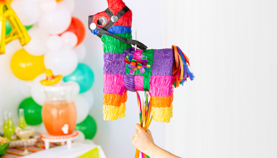 A child’s hand holding a rainbow-coloured jumbo donkey piñata with a variety of balloons and party snacks in the background.