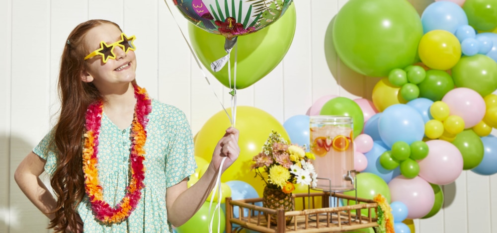 A girl wearing star sunglasses and an orange lei, holds a balloon bouquet in a yard filled with balloons and décor. 