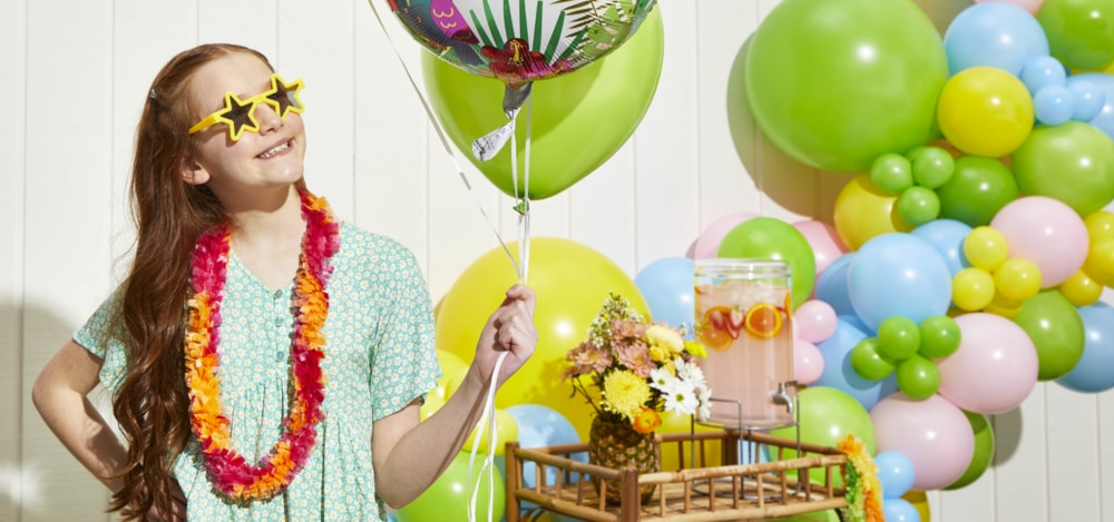 Une fille qui porte des lunettes de soleil en forme d’étoile et un collier de fleurs orange et qui tient un bouquet de ballons dans une cour remplie de ballons et de décorations. 