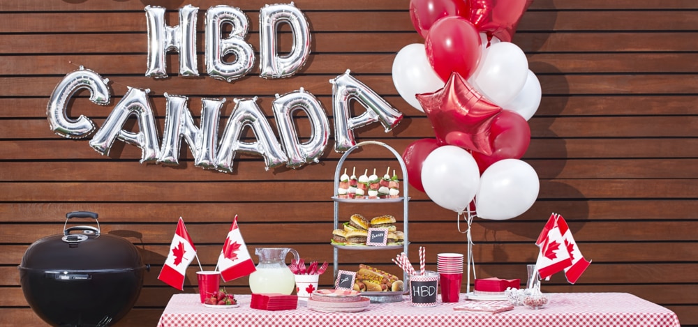 Un barbecue au thème de la fête du Canada décoré de drapeaux du Canada miniatures, de ballons rouges et blancs et d’une banderole de ballons argentés « HBD Canada. »