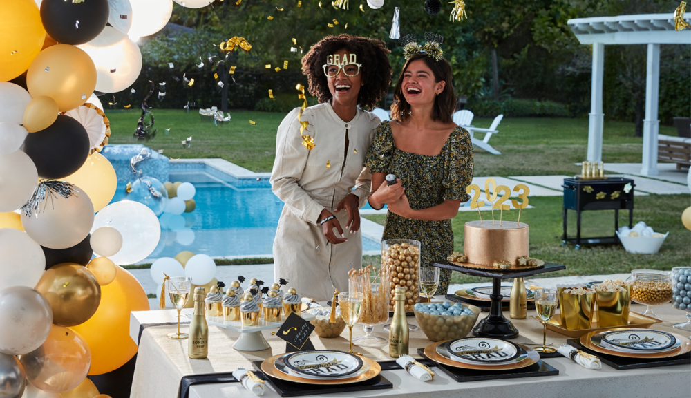 Two friends celebrating in a backyard party filled with various gold and black graduation-themed décor, balloons and party supplies. 