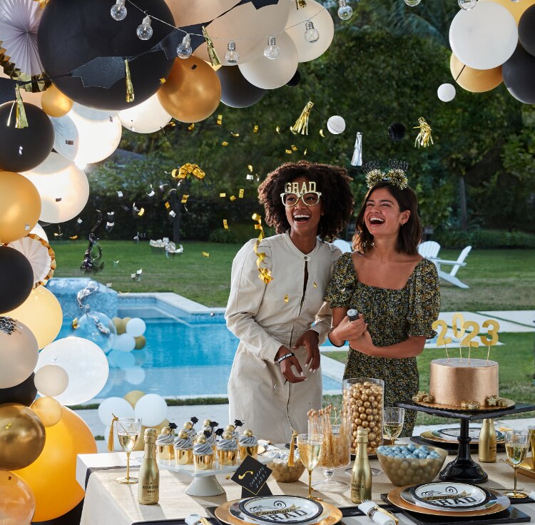 Two friends celebrating in a backyard party filled with various gold and black graduation-themed décor, balloons and party supplies. 