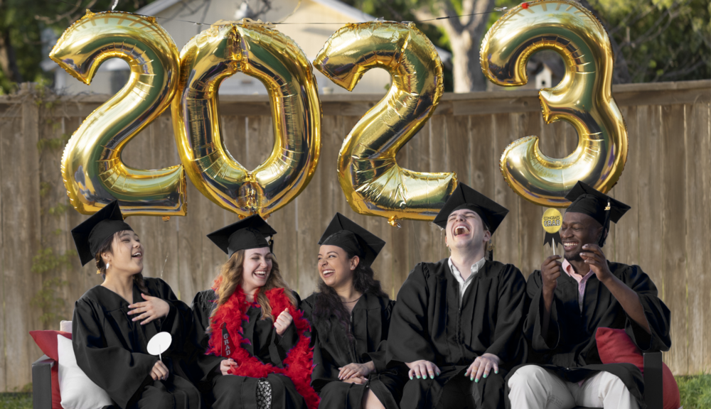 A group of graduates holding themed photo booth props sit in front of large helium-filled gold foil 2-0-2-3 number balloons.