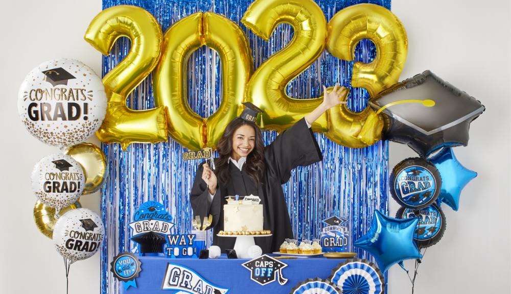 A woman wearing a graduation cap and gown posing at a table styled with graduation décor and balloon bouquets.