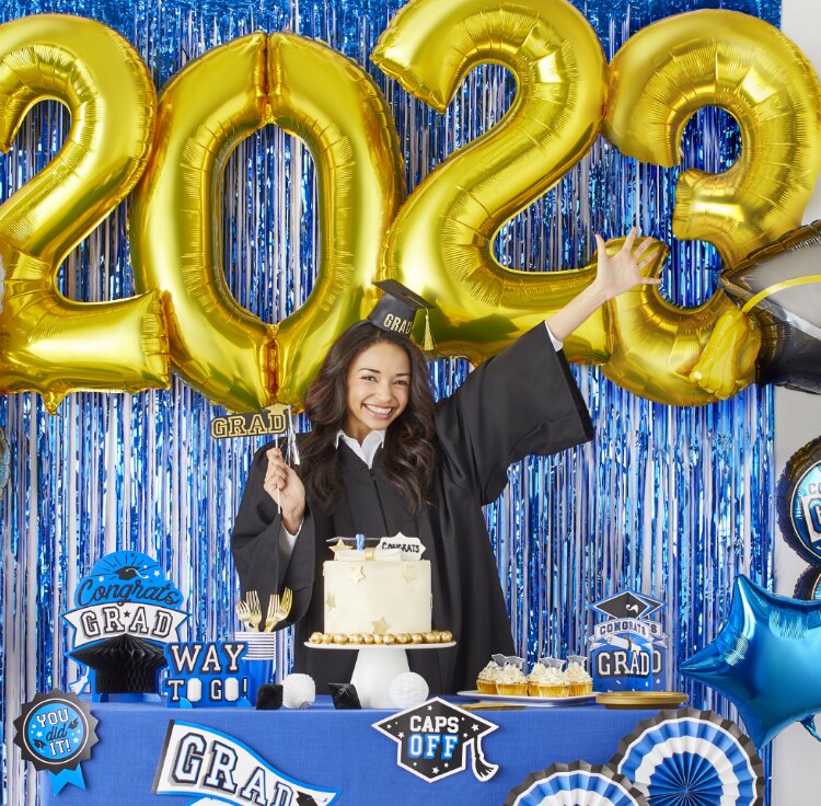 A woman wearing a graduation cap and gown posing at a table styled with graduation décor and balloon bouquets.
