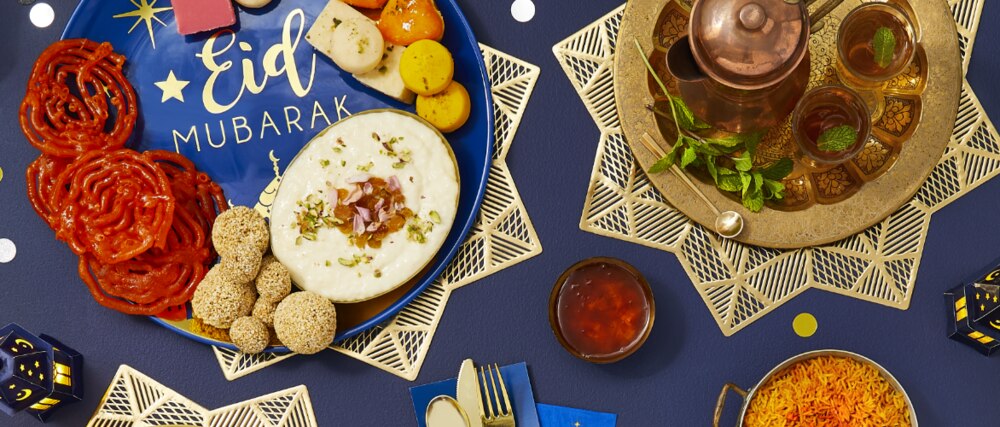 Various food items displayed on gold and blue Eid Mubarak-themed dinnerware and serveware.