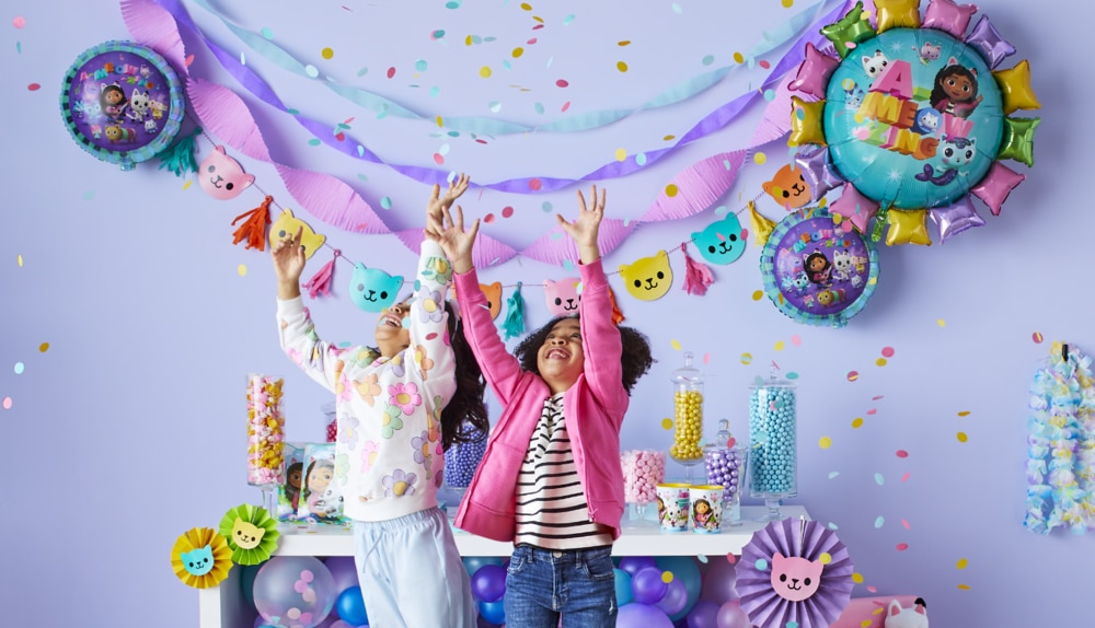 Two girls tossing confetti in a room decorated with colourful Gabby’s Playhouse themed foil balloons and decorations.