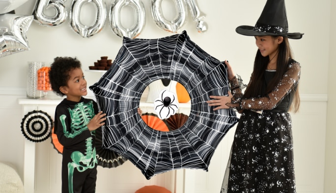 Two children dressed in Halloween costumes holding a large spiderweb-themed foil balloon.