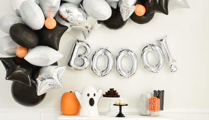 A silver “Booo!” balloon banner an orange, black and silver balloon arch hanging on a wall.
