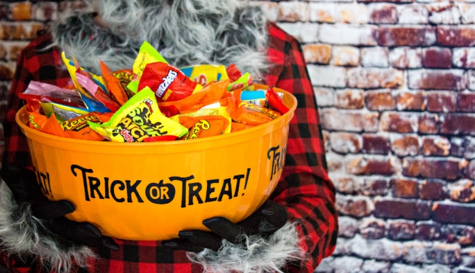 A person in a werewolf costume holding an orange “Trick-or-Treat” print bowl full of assorted packaged candies.