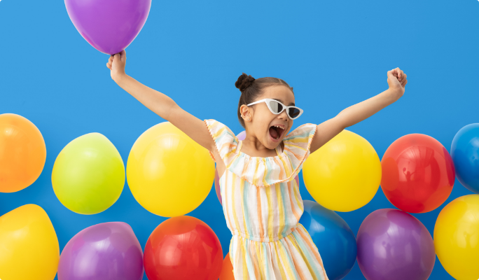 A girl holds a purple balloon while posing in front of various brightly coloured floating latex balloons.