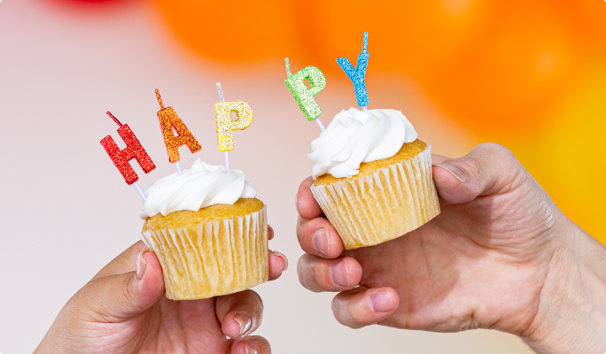 Two vanilla cupcakes with letter candles being held together to spell the word “happy.”
