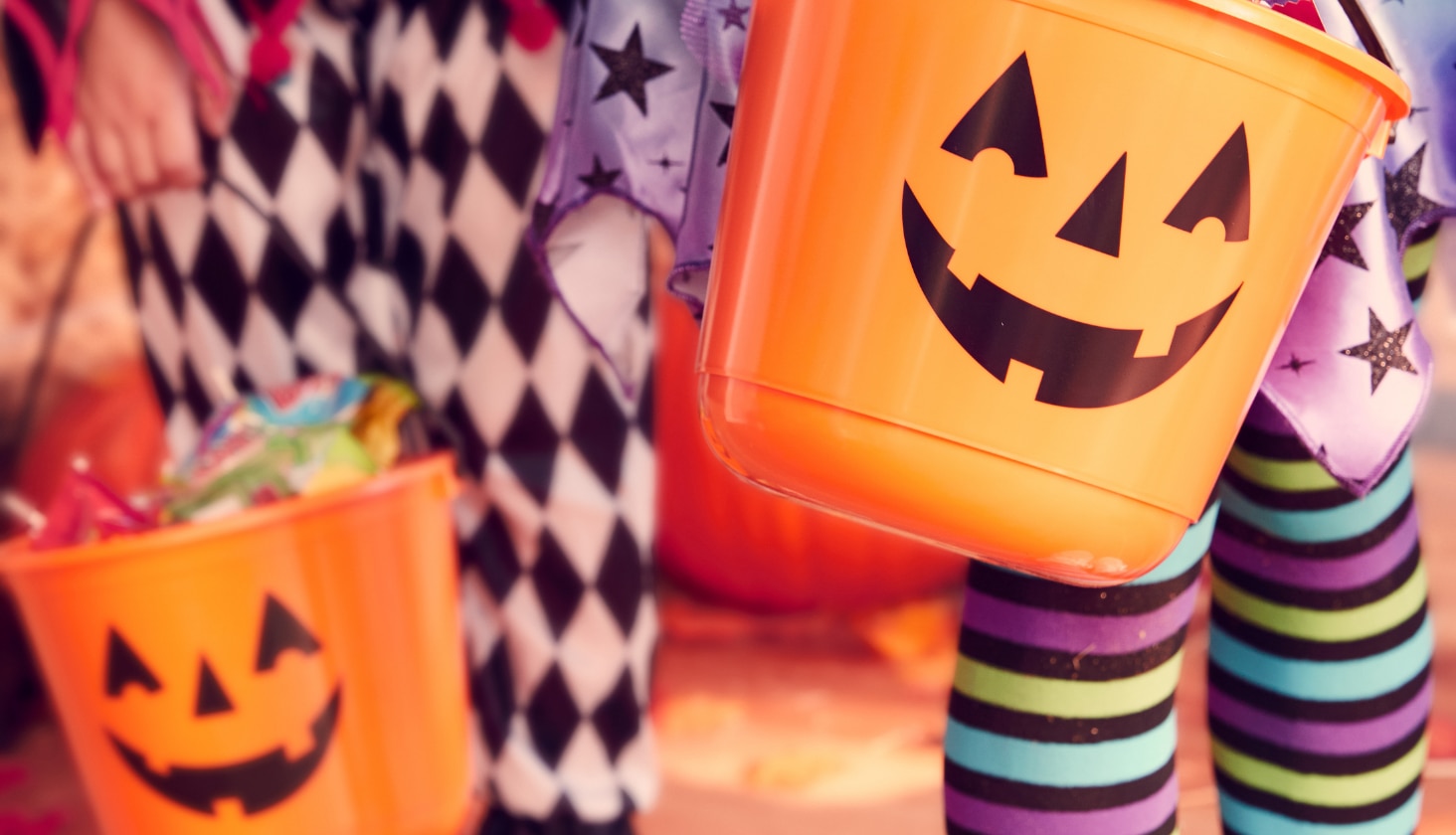 Two orange Jack-o’-lantern-themed treat pails being carried by children. 