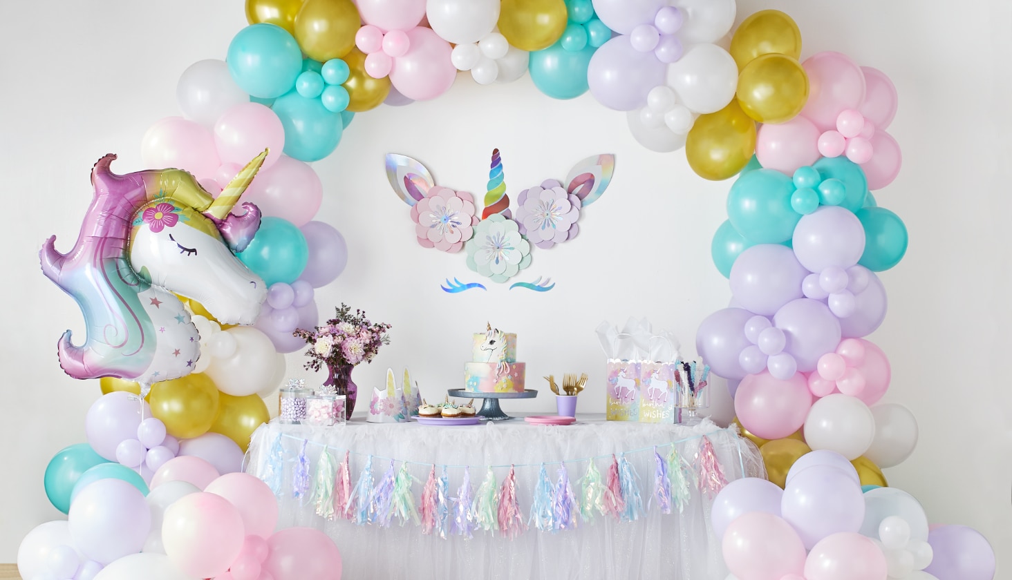 Une table à desserts décorée avec une guirlande de ballons, un ballon Licorne enchantée et plusieurs articles de fête thématiques.