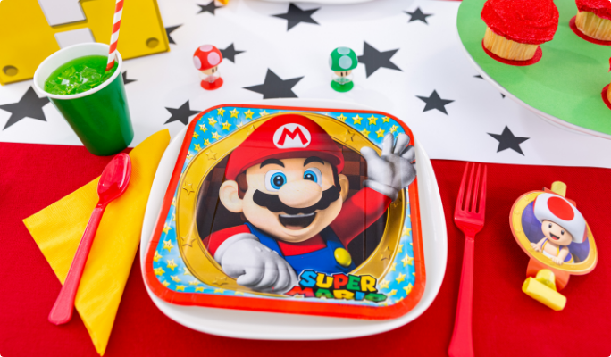 A square Super Mario paper plate and red plastic cutlery on table with various party supplies. 