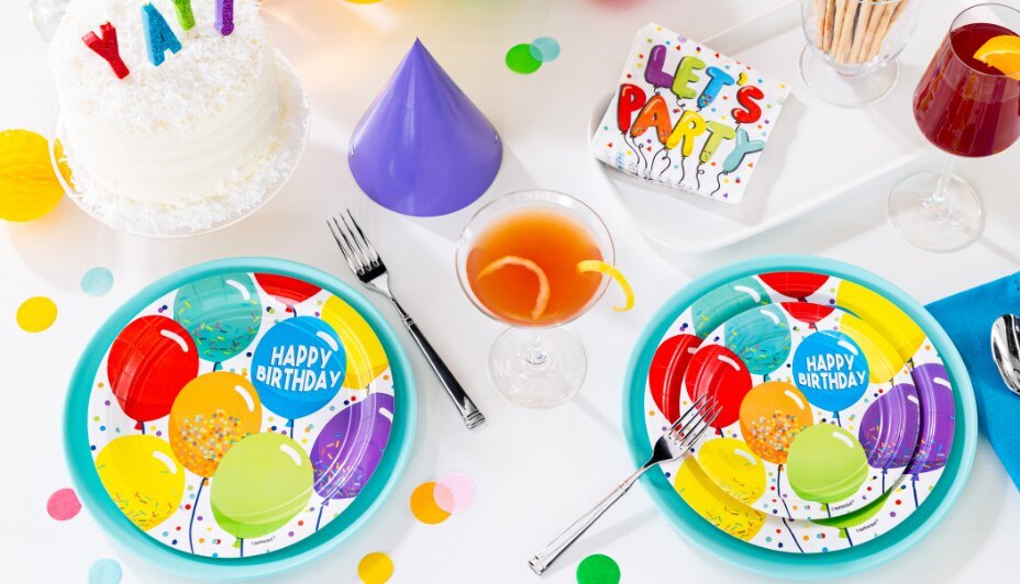 A table set with multicoloured balloon birthday celebration tableware, cake and confetti.