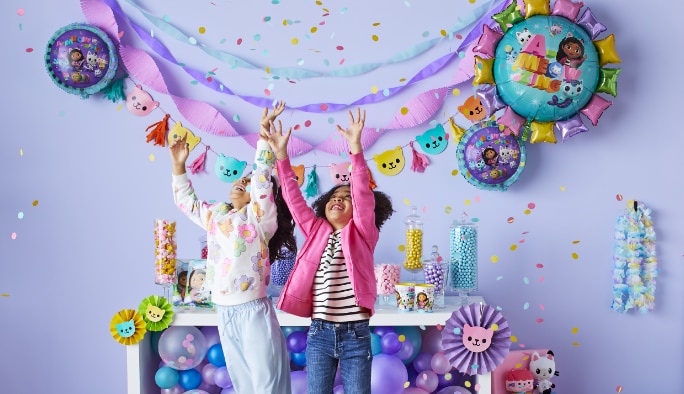 Two young girls throwing confetti into the air in front of a birthday decor arrangement.