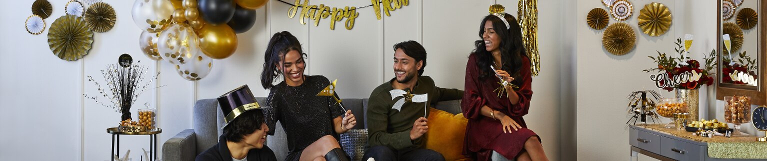 Friends dressed for New Year's Eve celebrations, sitting in a living room decorated in black, white and gold decorations, all holding noisemakers.
