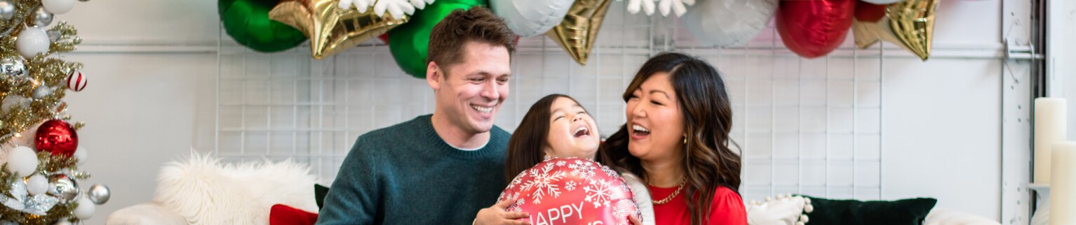 A smiling mom, dad and young daugher holding a balloon that reads "Happy Holidays" all sitting on a white couch surrounded by Christmas decorations.