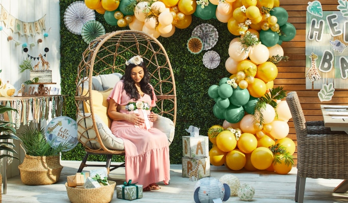 A pregnant woman sitting on a patio decorated with a yellow and green balloon arch, a “hello baby” banner and various jungle-themed decor.