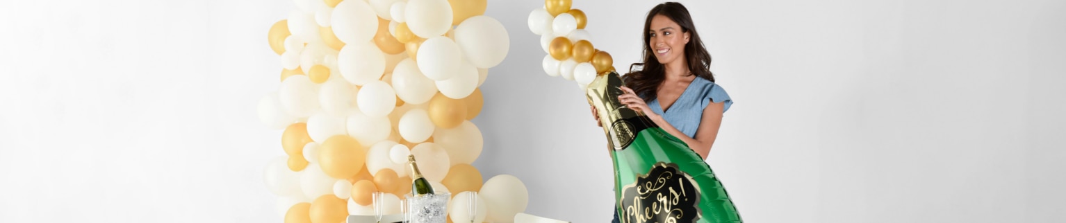 A woman holding a large bubbly champagne-shaped balloon with white and gold balloons and a set table in the background.