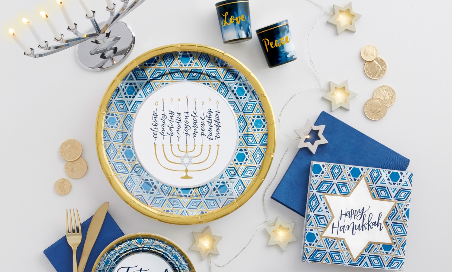 A menorah, plate, napkins, cups, cutlery, coins and star string lights on a white table.