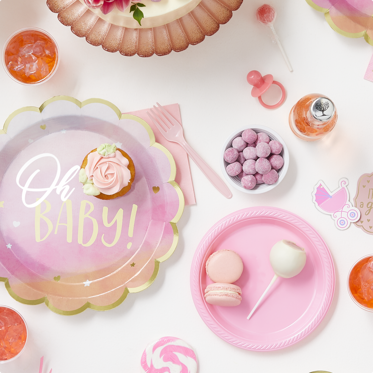 An "Oh Baby" metallic gold and pink plate, a pink round paper plate, pink cutlery, and an assortment of pink dessert on a white backdrop.