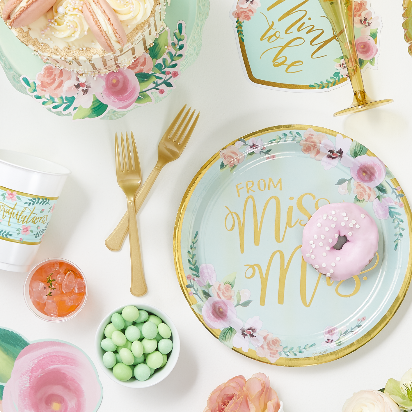 A round paper plate featuring pink flowers and a gold border and a "From Miss to Mrs" headline, gold cutlery and cups and an assortment of pink and mint dessert on a white backdrop.