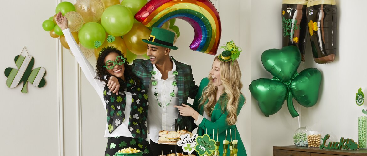 Three people dressed in St. Patrick's Day wearables appear to be having fun in a room decroated with themed green, gold, rainbow and shamrock-shaped balloons.