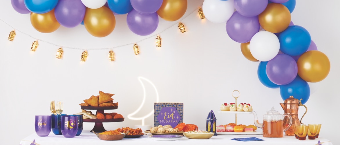 Hanging decorative lanterns and purple, gold, blue and white balloons hang above a table decorated with Ramadan & Eid tableware.