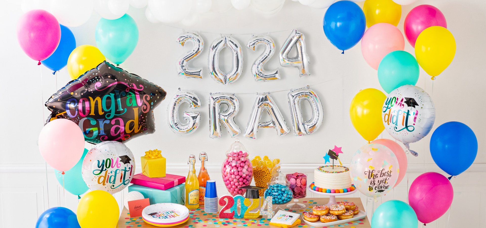 A dessert table surrounded by a multi-coloured balloon bouquet, silver letter and number balloons that spell out 2024 GRAD and various graduation party decorations.