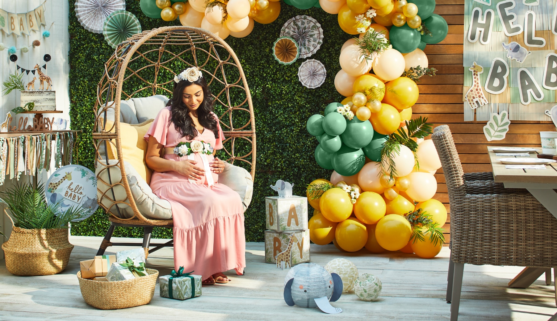 Une femme enceinte tenant son ventre assise sur une chaise sur une terrasse décorée d’une arche de ballons jaunes et verts, d’une banderole « Hello Baby », et de décorations à thème de jungle.