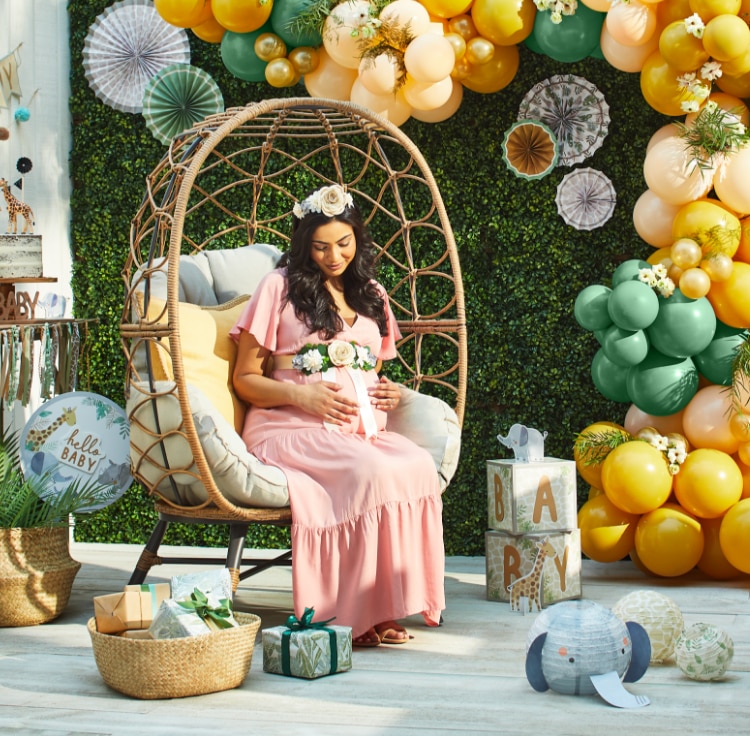 A pregnant woman sitting in a chair holding her belly, on a patio decorated with a yellow and green balloon arch, a “hello baby” banner and various jungle-themed decor.