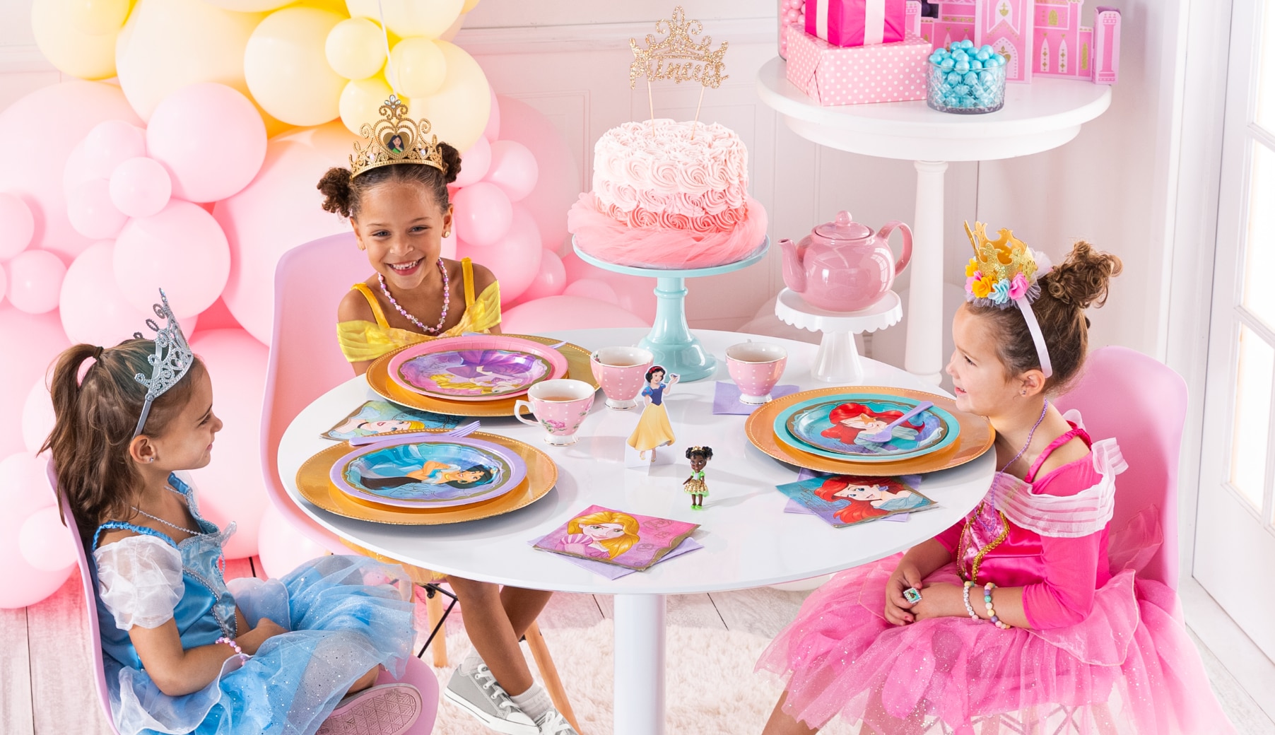 Trois jeunes filles déguisées en princesses assises à une table de desserts d’anniversaire avec des assiettes, des serviettes de table et des tasses à thé à thème de princesses.