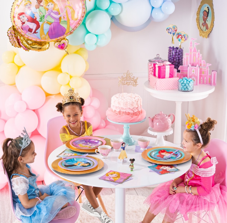 Trois jeunes filles déguisées en princesses assises à une table de desserts d’anniversaire avec des assiettes, des serviettes de table et des tasses à thé à thème de princesses.